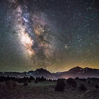 The milky way above a foggy landscape with mountains in the background and a forest in the foreground.