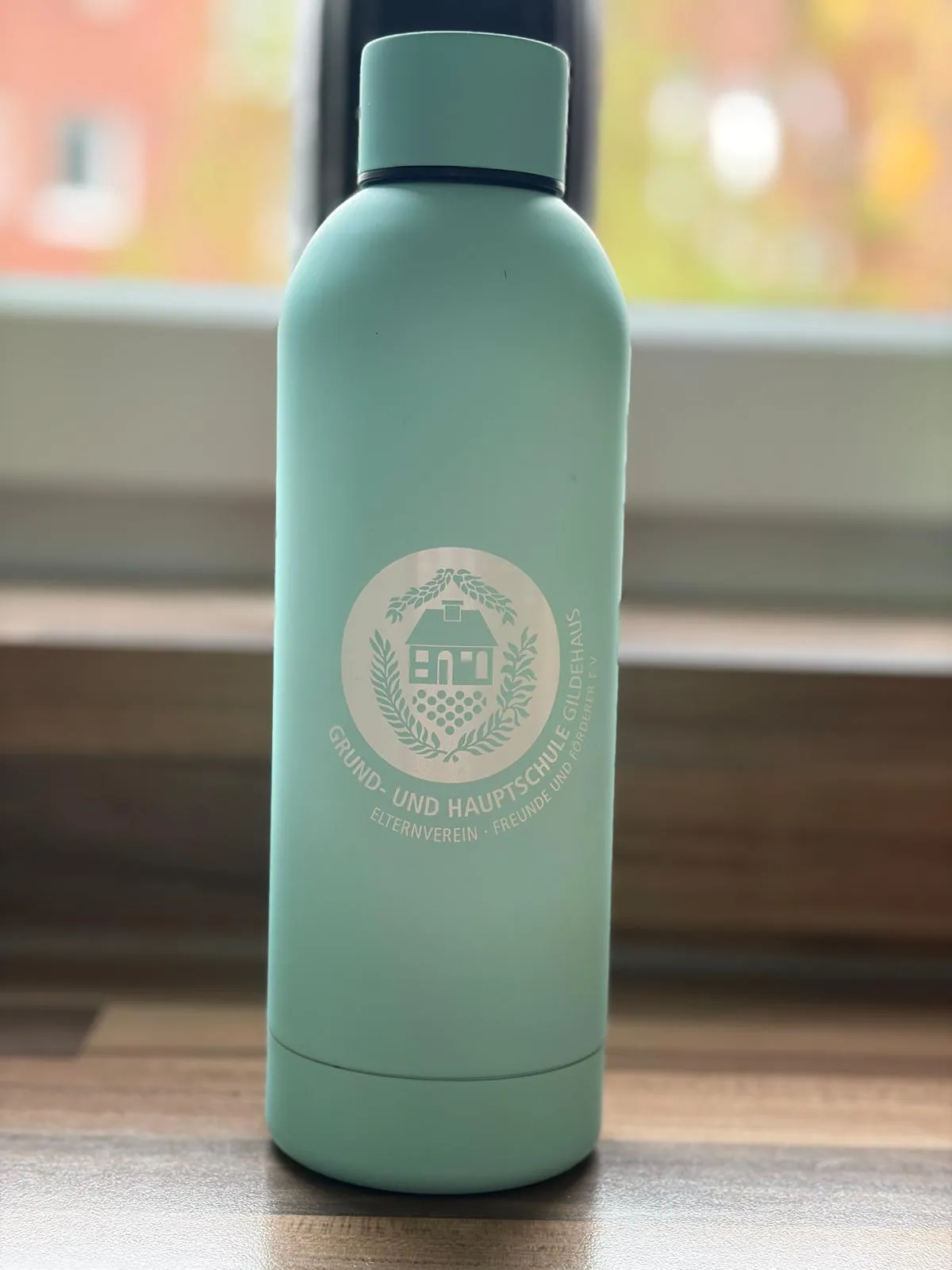 Neue mintgrüne Trinkflasche mit eingraviertem Logo des Elternvereins der Grund- und Hauptschule Gildehaus auf einer Fensterbank.