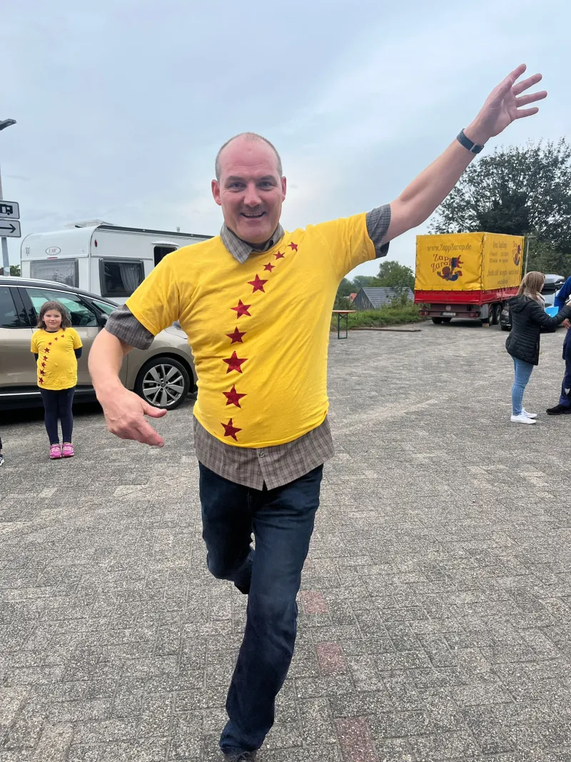 Ein Helfer im gelben Zirkus-T-Shirt mit roten Sternen läuft mit ausgebreiteten Armen über den Platz vor dem Zirkuswagen.