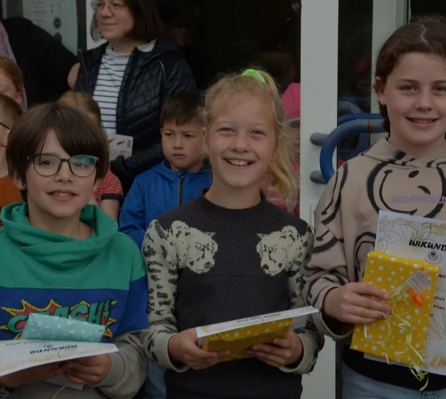 Drei Kinder halten nach dem Vorlesewettbewerb ihre Urkunden und in gelbes Papier eingepackte Buchgeschenke in den Händen.
