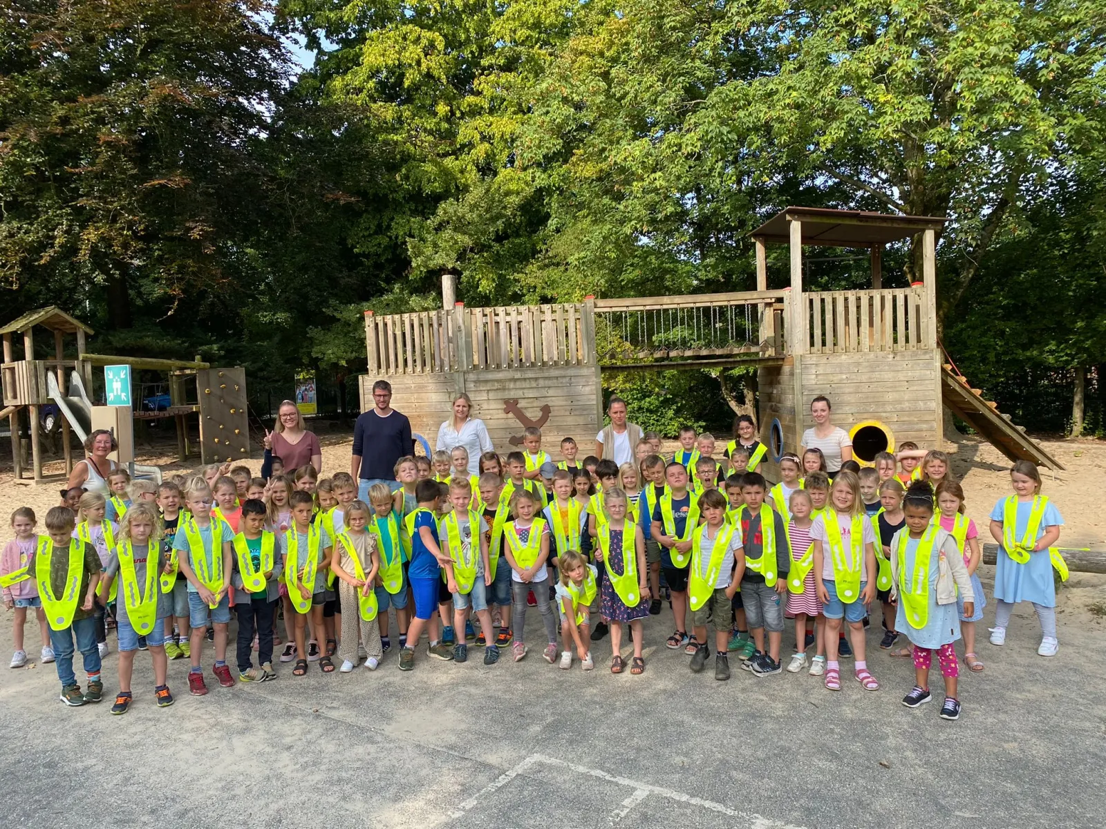 Alle Erstklässler stehen mit ihren neuen gelben Warnwesten vor dem Klettergerüst auf dem Schulhof, dahinter die Lehrkräfte.